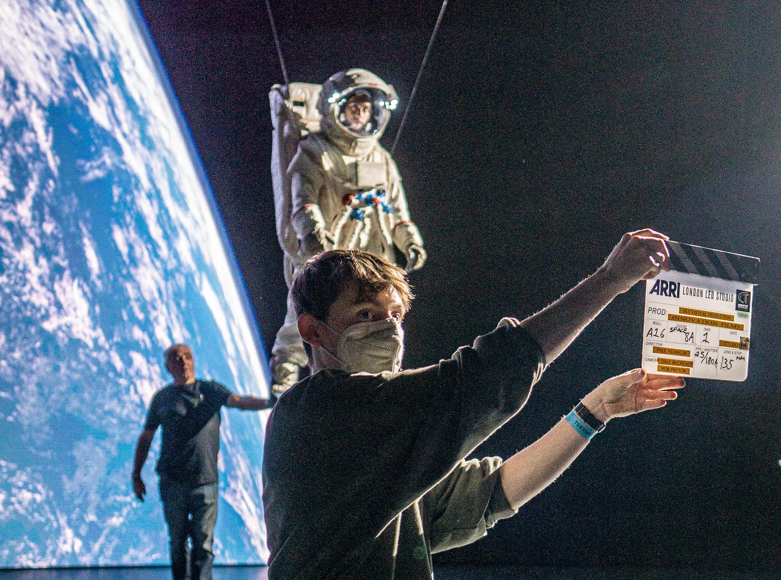 Charlie Hedger holding a clapper board. Behind him is an astronout suspended on wires with an LED image of the earth behind.