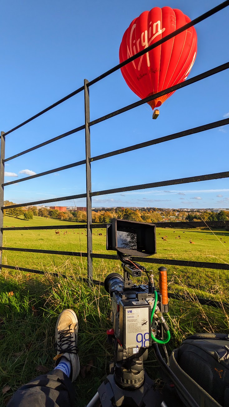 POV shot of Charlie Hedger sat in a field with a cinema camera. In the distance there are Deer and a hot air balloon floats above.