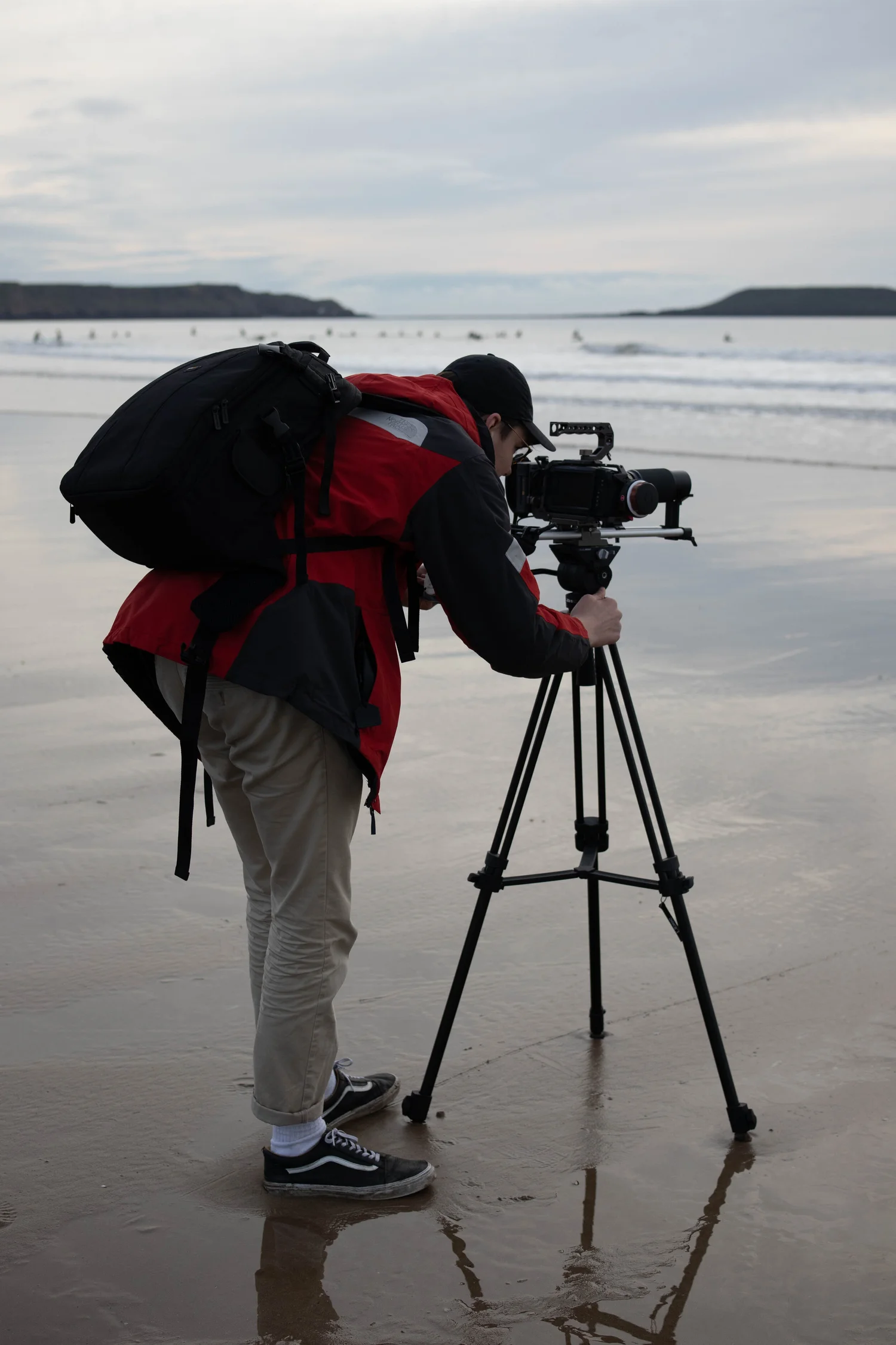 A camera operator on a beach. He is leant over a camera facing out to sea. Surfers are behind him in the distance.