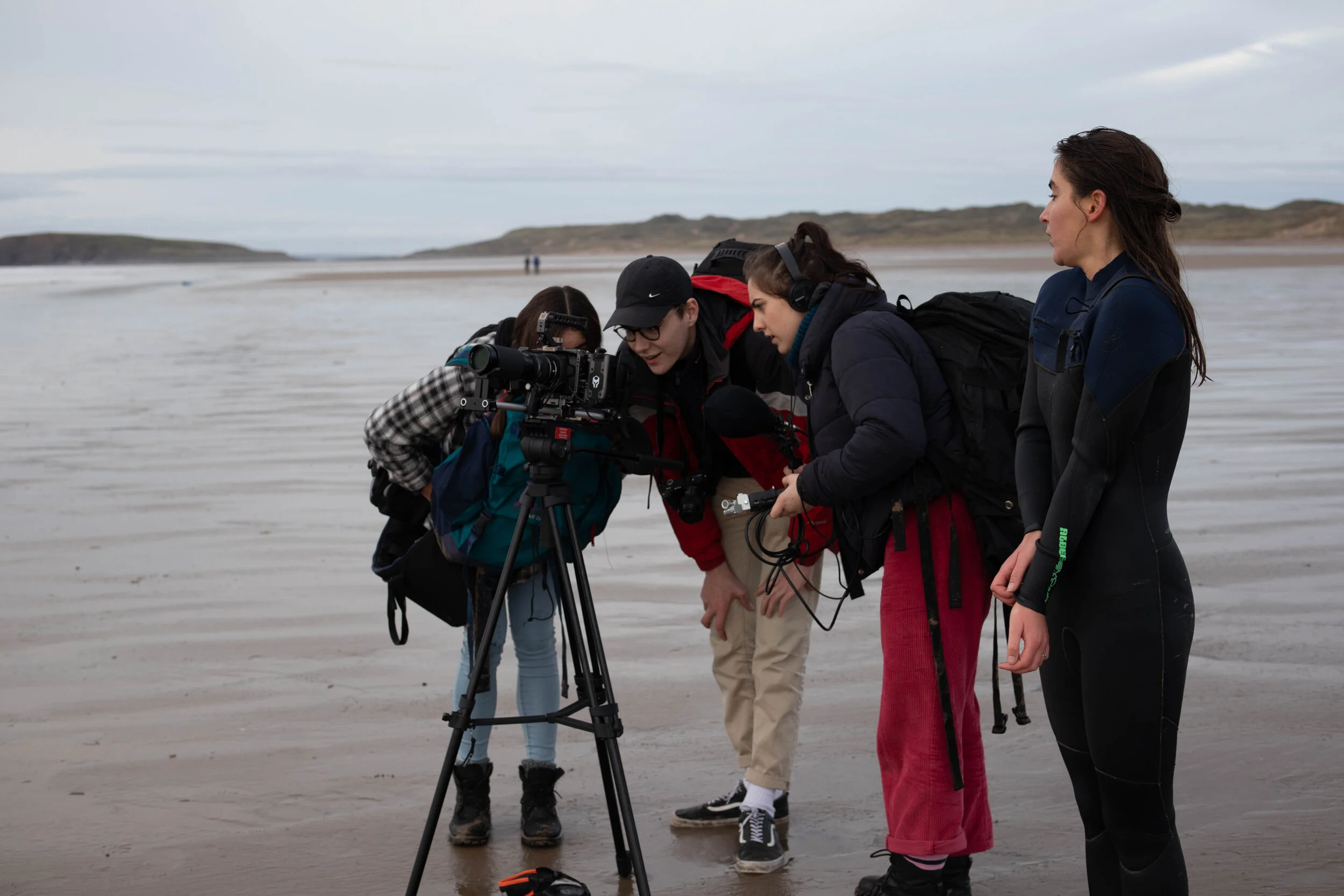 A group of filmmakers gather arounda a camera on a beach. The camera is on a tripod facing out to sea.