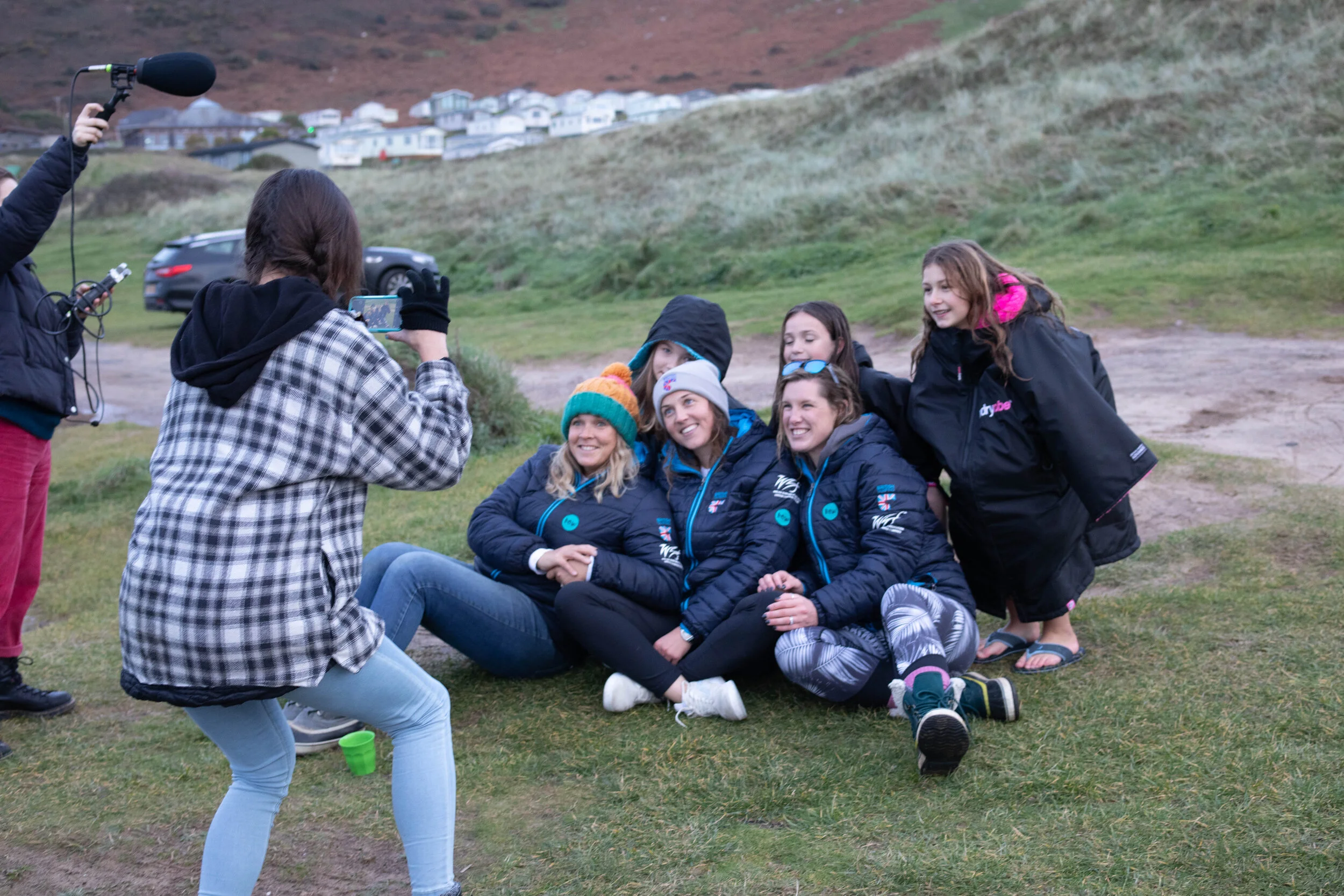 A group of female surfres are having their photo taken on a patch of grass in front of some sand dunes.
