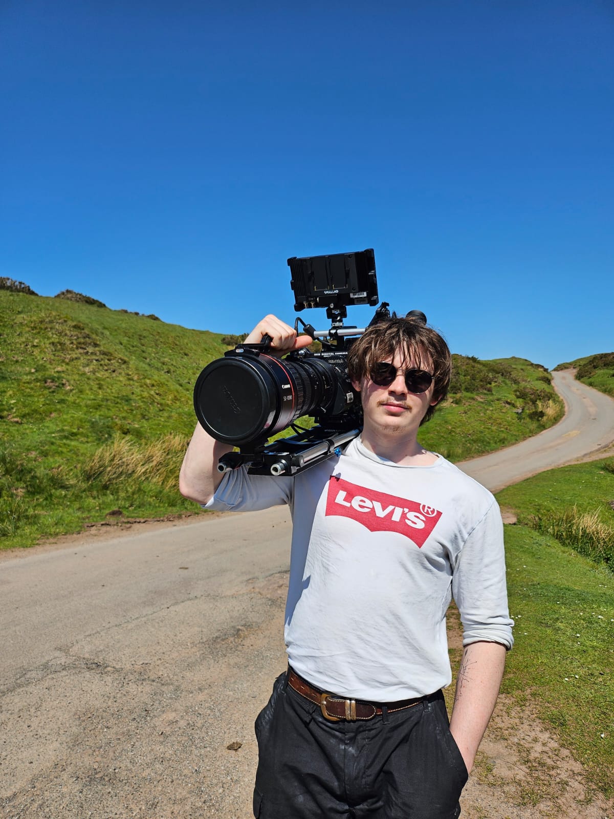 Charlie Hedger holidng a large cinema camera with a Canon CN20 Zoom Lens on it. He is on the top of a hill. Behind him is a winding road.