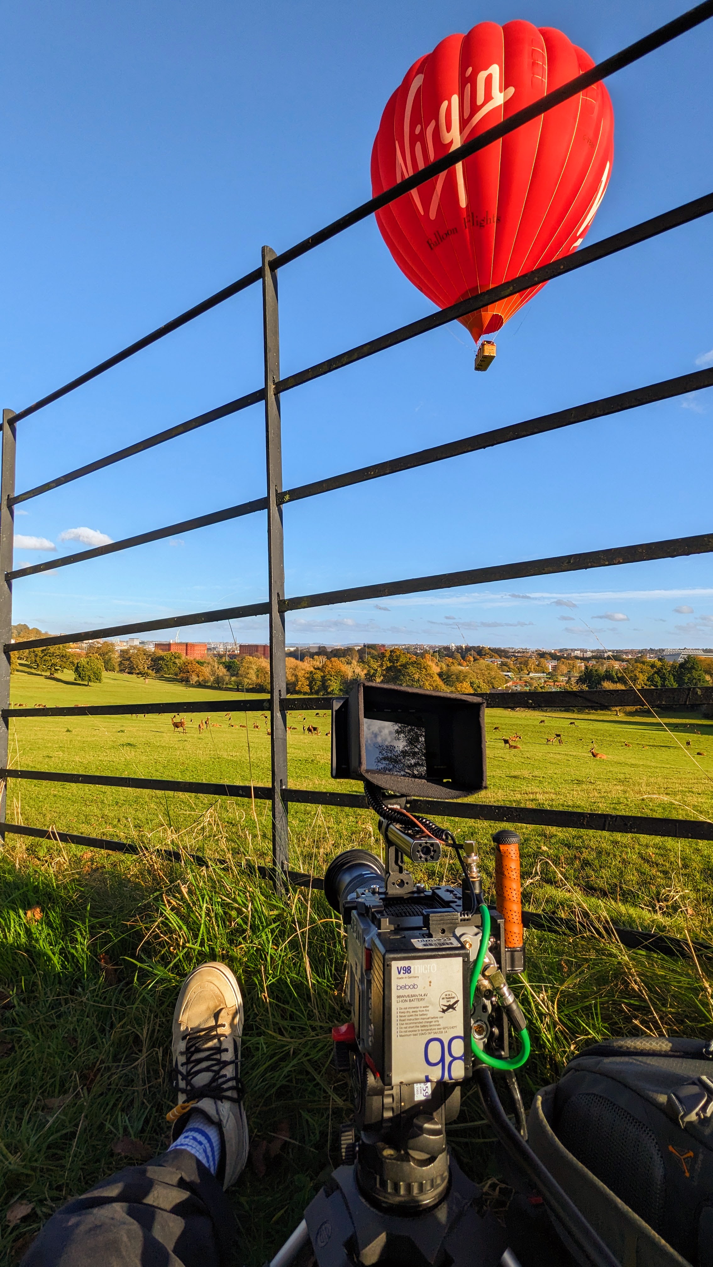 POV shot of Charlie Hedger sat in a field with a cinema camera. In the distance there are Deer and a hot air balloon floats above.