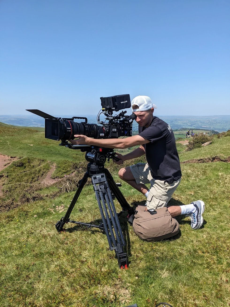 A young camera operator on the top of a hill. He holds a large cinema camera on a tripod.