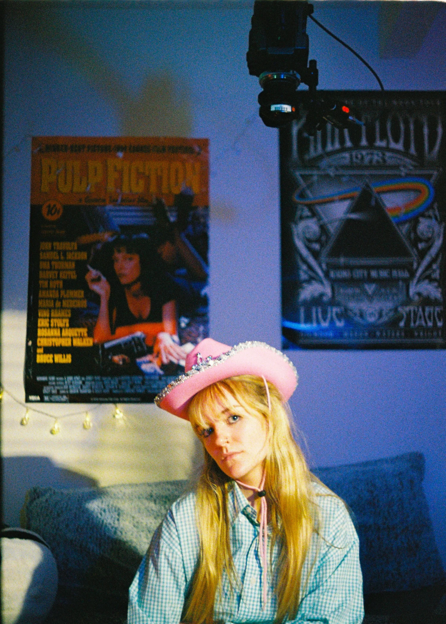A girl sits on her bed with a pink cowboy hat on. Behind her are movie poster and above you can see a cinema camera looking down on her.