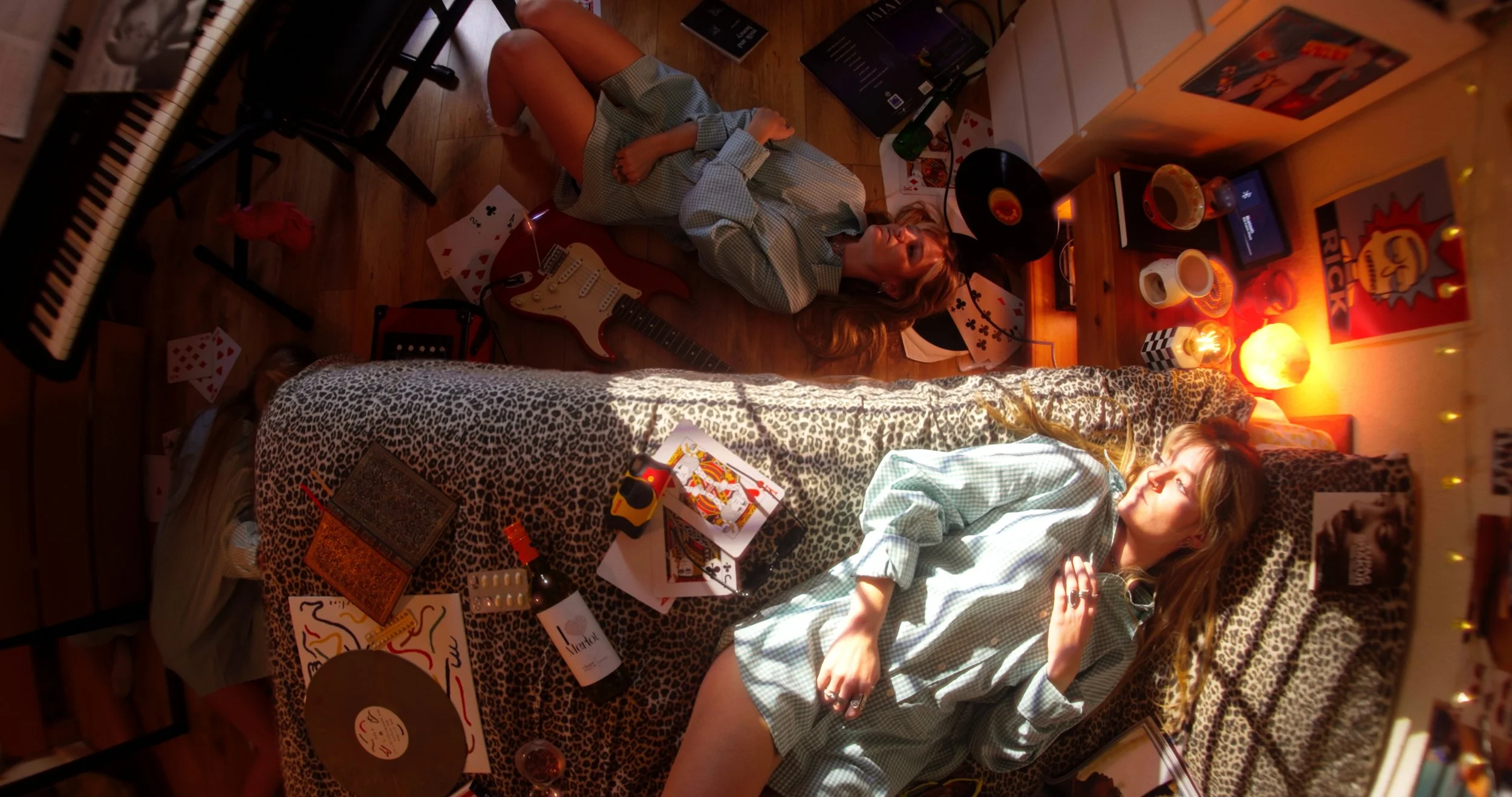 A girl is lying on a messy bed. Below her a clone of the same girl is lying on the floor next to her guitar.
