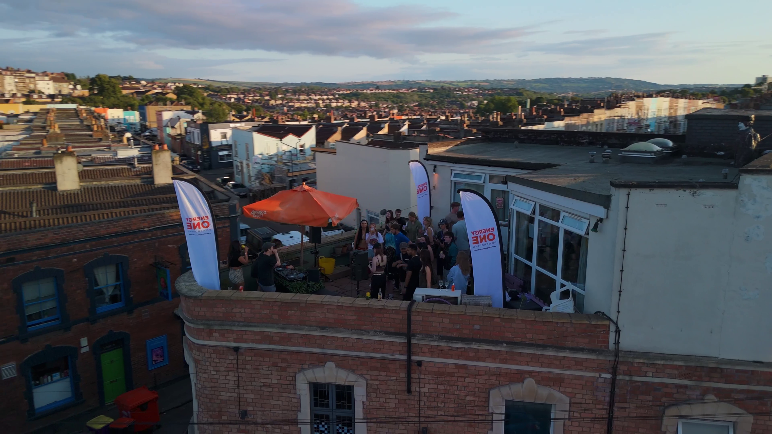 An aerial shot of a group of people on a roof terrace. They are dancing in front of a set of DJ Decks.