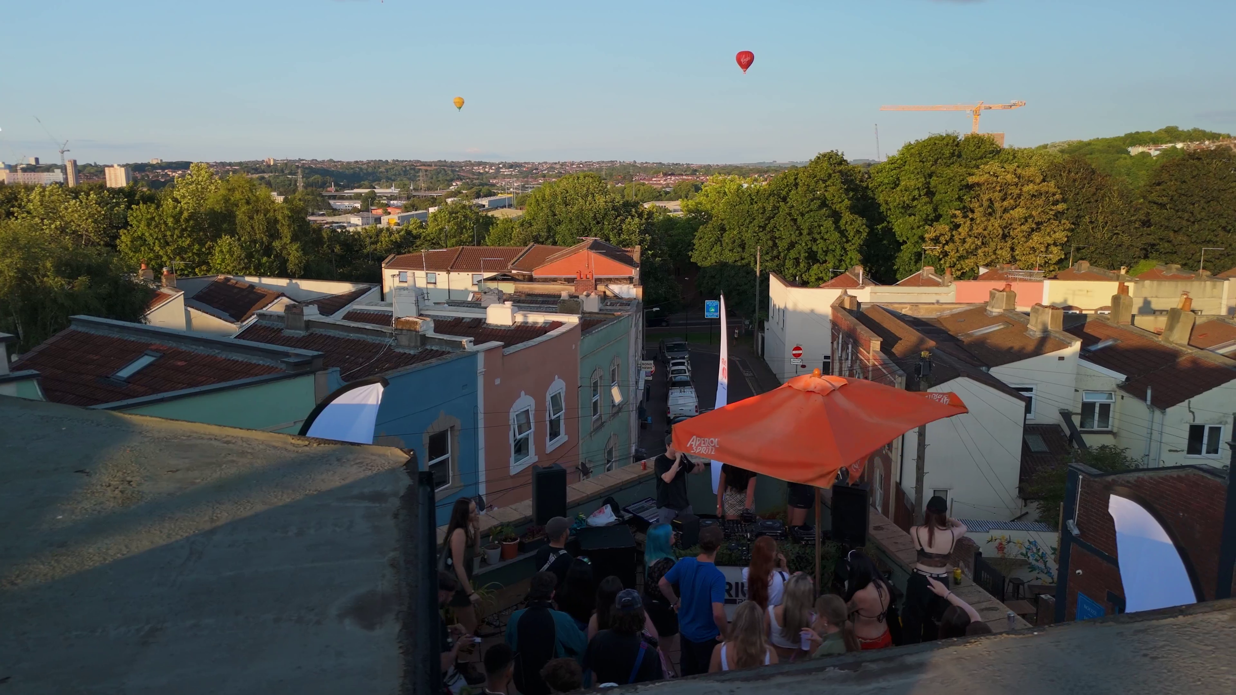 An aerial shot of a group of people and DJ decks on a roof terrace. Behind them you can see hot air balloons in the sky.