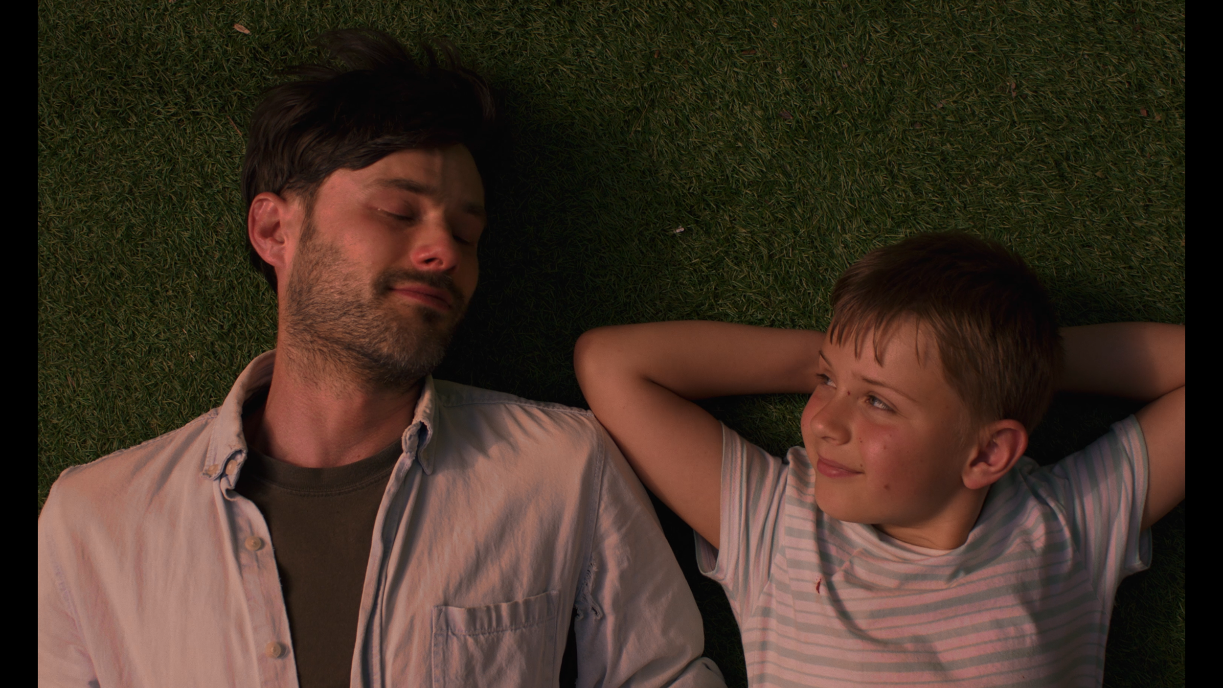 A father and son lie together on the grass. The fathers eyes are closed. The son looks at the father. Both are happy.