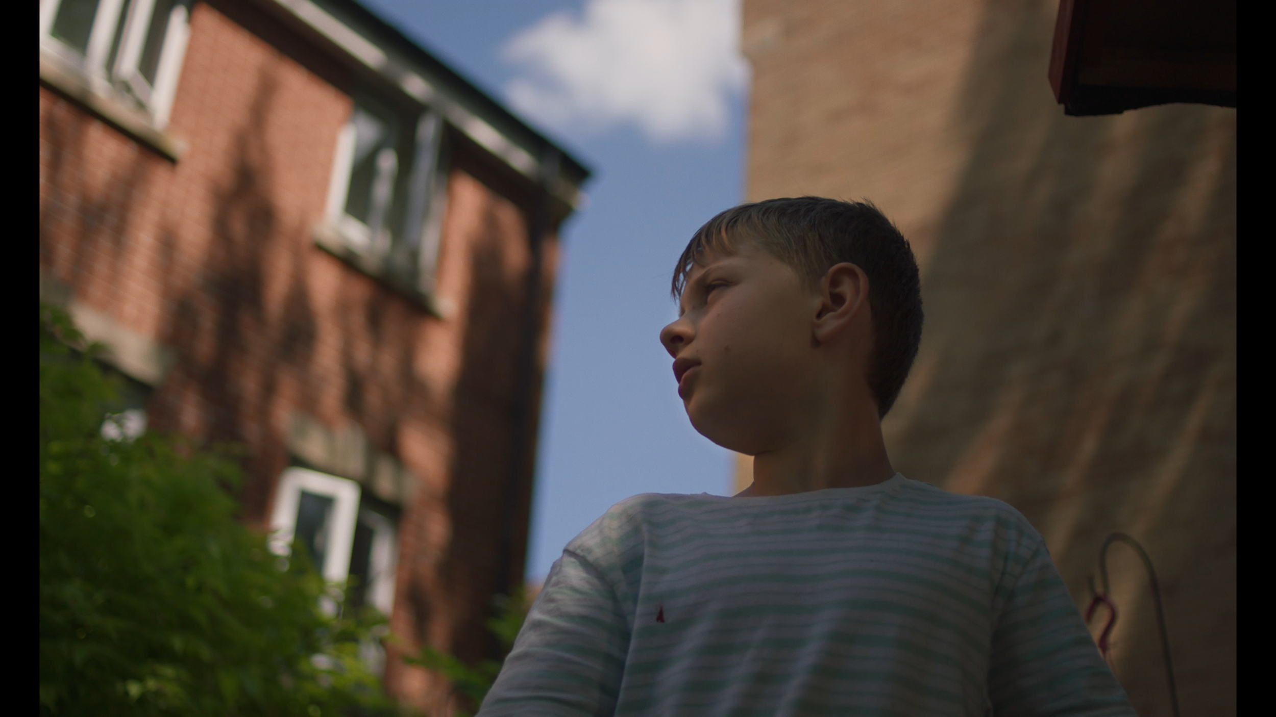 A young boy is looking off camera. He is wearing a stripey shirt and is outdoors in a garden.